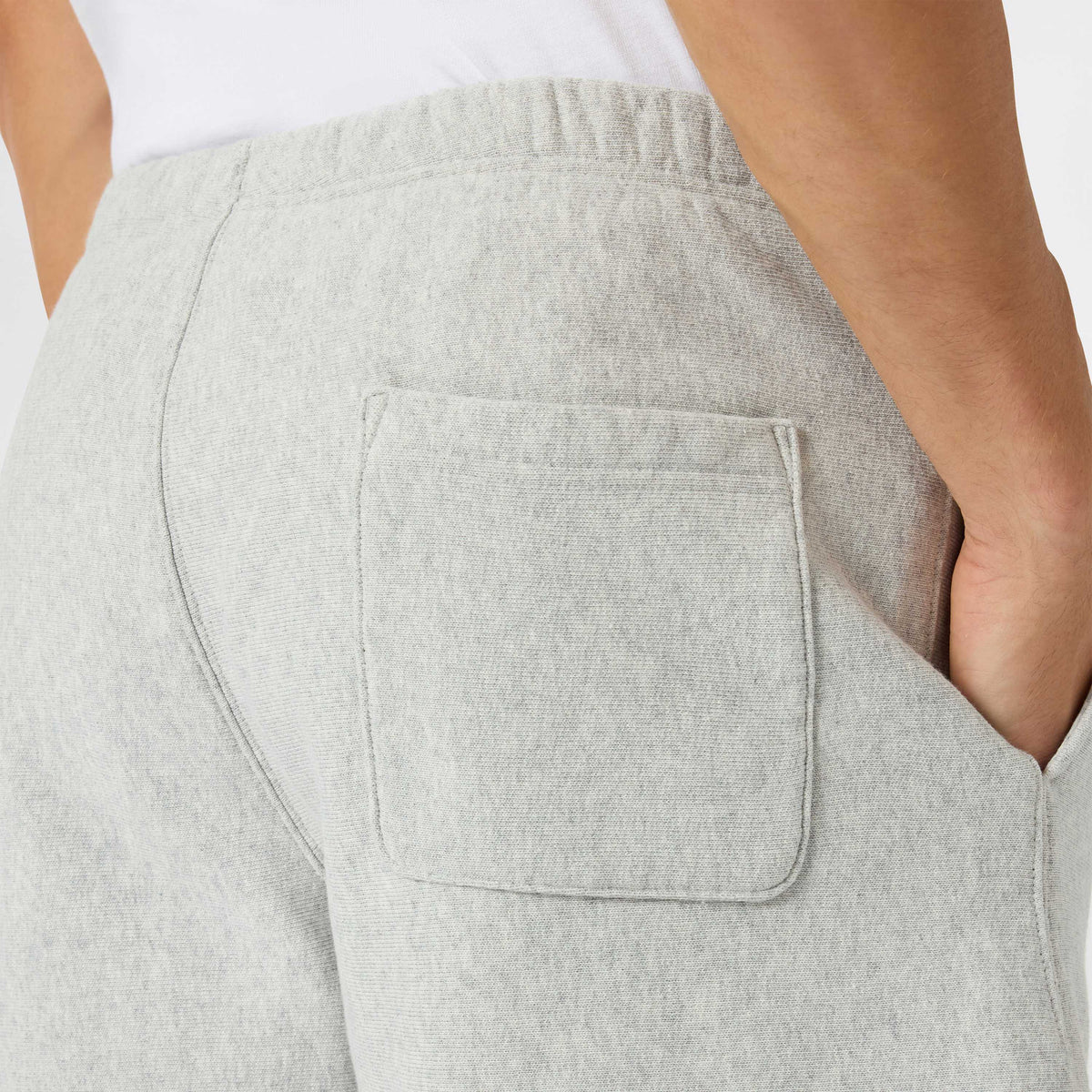 Close-up of a person wearing Champion Men’s Knitted Shorts in light gray, with one hand in the side pocket. Inspired by classic men’s sportswear style, the shorts feature a visible back patch pocket and an elastic waistband. The person is also wearing a white shirt.