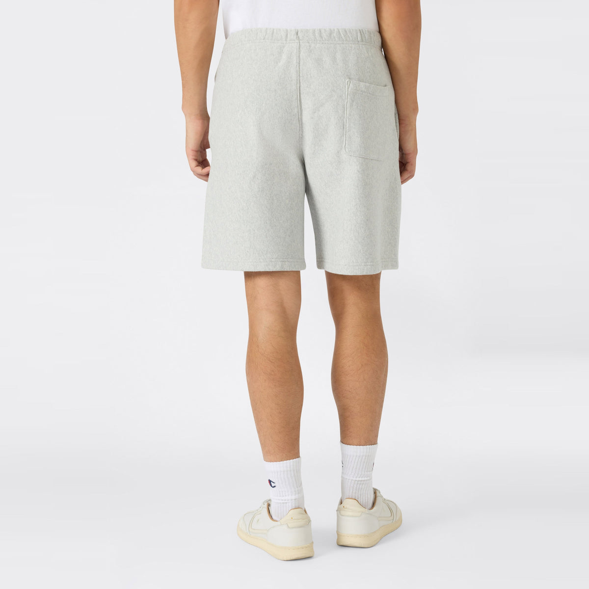 A person is seen from behind, standing against a plain white background, wearing light gray Champion Men’s Knitted Shorts crafted from heavyweight fleece. The sportswear-inspired shorts feature a single back pocket on the right side and are paired with white socks and cream sneakers.
