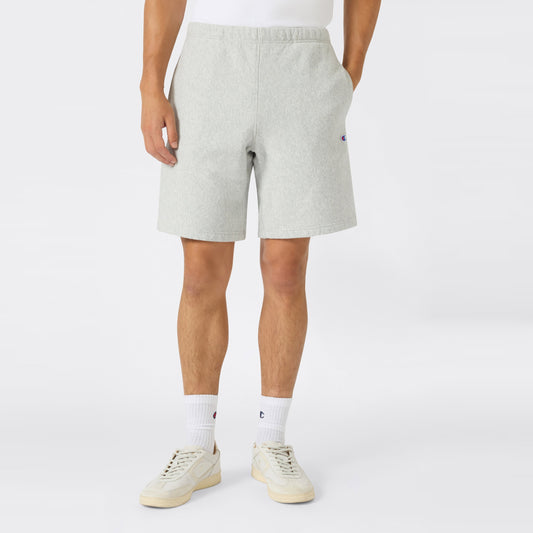 A person wearing Men’s Knitted Shorts by Champion in light gray, paired with white socks and cream sneakers, stands against a plain background. Their hands rest in their pockets, and only the lower half of their body is visible—showcasing casual men’s sportswear from Champion crafted from heavyweight fleece.