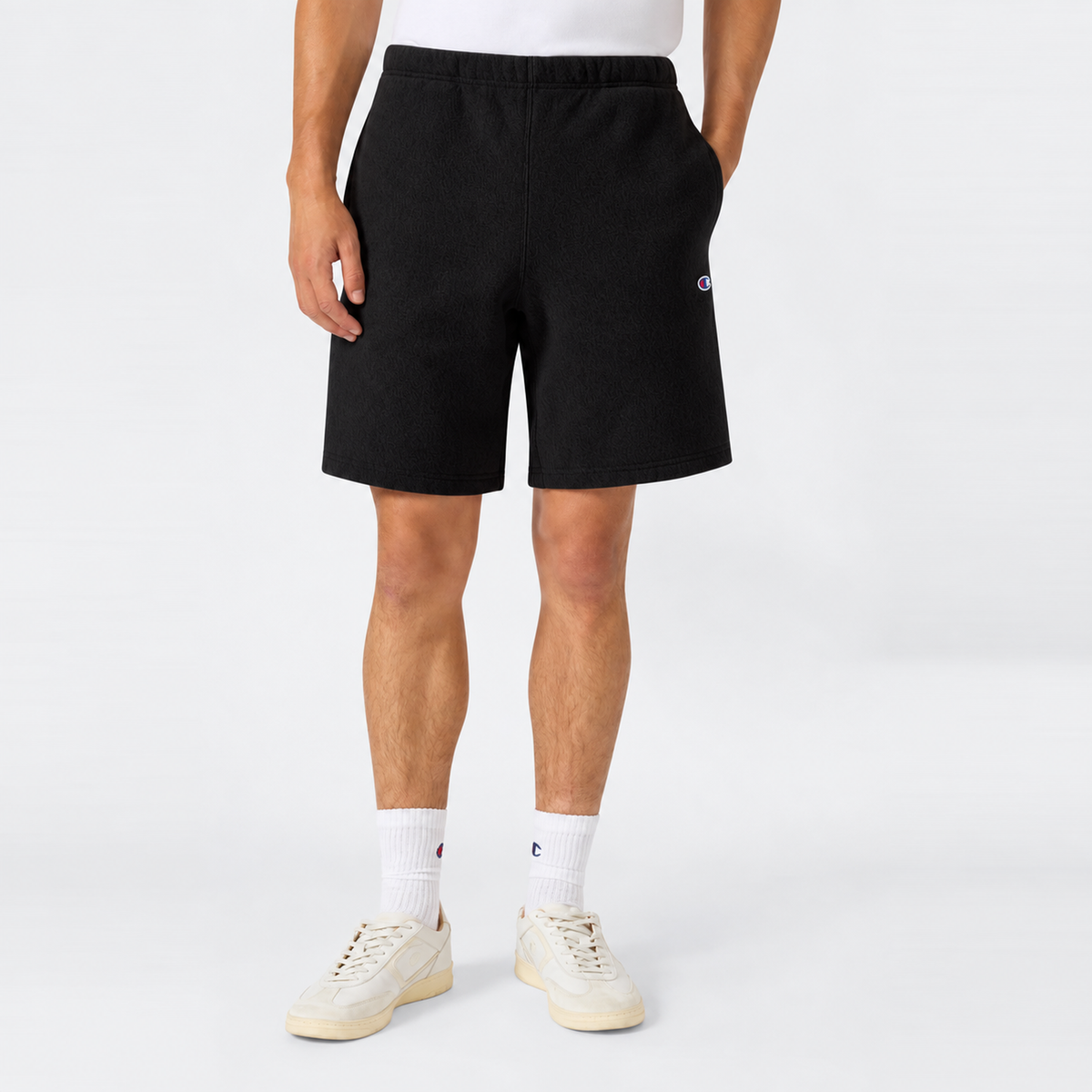 A person wearing black Champion Men’s Knitted Shorts, paired with a white t-shirt, white socks, and white sneakers, stands against a plain light background. Only the lower half of their body is visible, highlighting classic men’s sportswear from Champion.