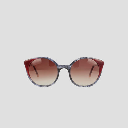 The Champion Women's Polarized Flirty Round sunglasses feature gradient brown polarized lenses, marbled gray handmade acetate frames, and solid dark red accents on the upper corners, shown against a plain light gray background.