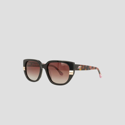 Champion Women’s Polarized Modified Cateye sunglasses featuring black acetate frames with gold accents along the sides and brown gradient polarized lenses. The handmade design includes tortoiseshell-patterned arms adorned with a discreet logo and finished with a pink tip, all presented against a plain light gray background—a stand-out piece of luxury eyewear.