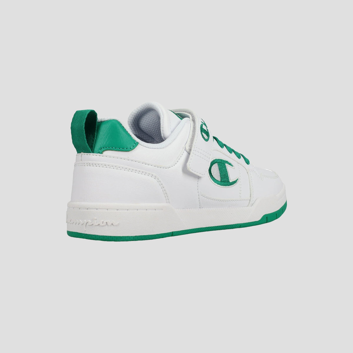 The Champion Kids' Arena Power Low sneakers are white with green accents on the heel, sole, and logo. Viewed from a rear side angle, they feature a back pull tab and a hook-and-loop strap over the laces for easy wear.