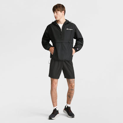 A person stands against a white background, wearing a black Champion jacket, Champion 7" Woven Sport Shorts, white socks, and black sneakers. They have tattoos on one leg and look to the side with hands in their jacket pockets.
