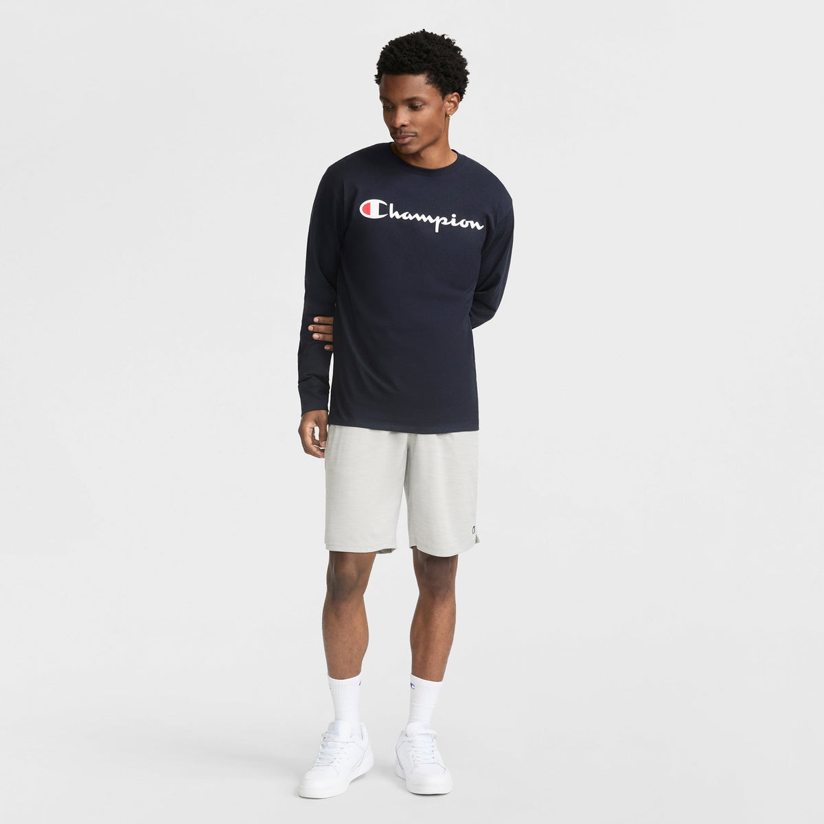 A man stands against a white background wearing a navy Champion long-sleeve, 10" Moisture Wicking Sport Shorts by Champion in light gray, white crew socks, and white sneakers. He gazes left with one hand down and the other holding his shorts.