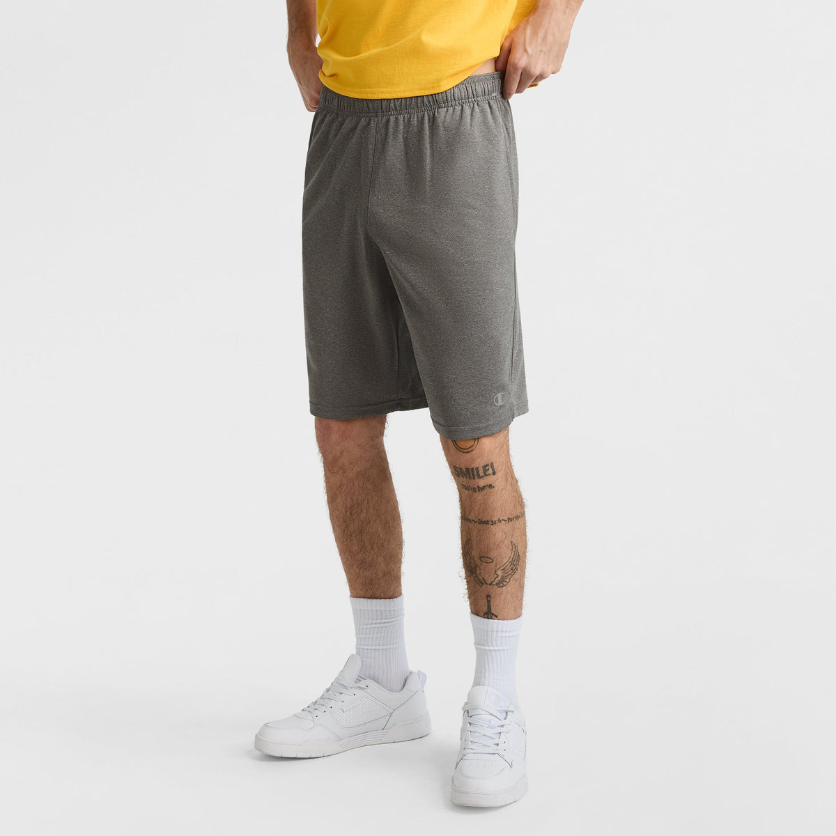 A person stands on a plain white background wearing a yellow shirt and Champion 10" Moisture Wicking, Sport Shorts for men. Their hands are in their pockets, tattoos cover their lower legs, and they complete the look with white socks and sneakers.