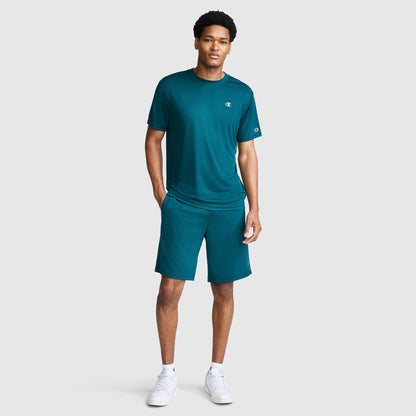 A man wearing a teal athletic t-shirt and Champion 10″ Moisture Wicking Sport Shorts stands with one hand in his pocket, looking forward. He is also wearing white sneakers against a plain, light gray background.