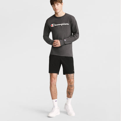 A man with tattoos on his left leg and brown, bang-styled hair stands against a plain background wearing a dark gray Champion long-sleeve shirt and Champion 10" Moisture Wicking Sport Shorts, paired with white socks and white sneakers.