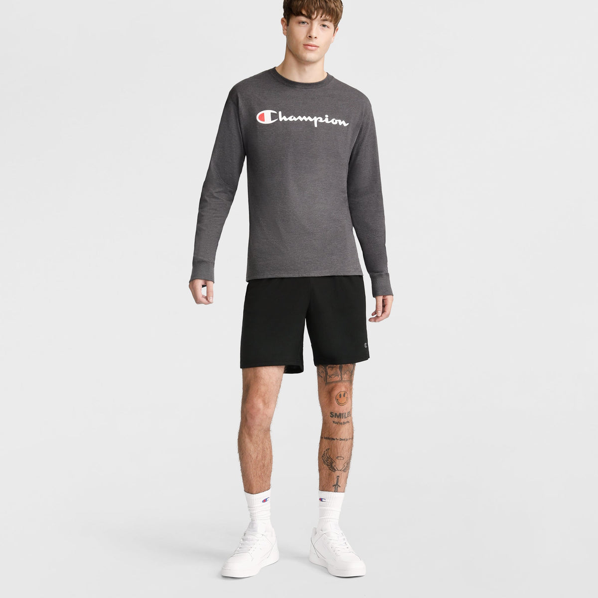 A young man with tattoos on his right leg wears Champion 10" Moisture Wicking Sport Shorts, a gray long-sleeve Champion shirt, white socks, and white sneakers against a plain background, showing a neutral expression.
