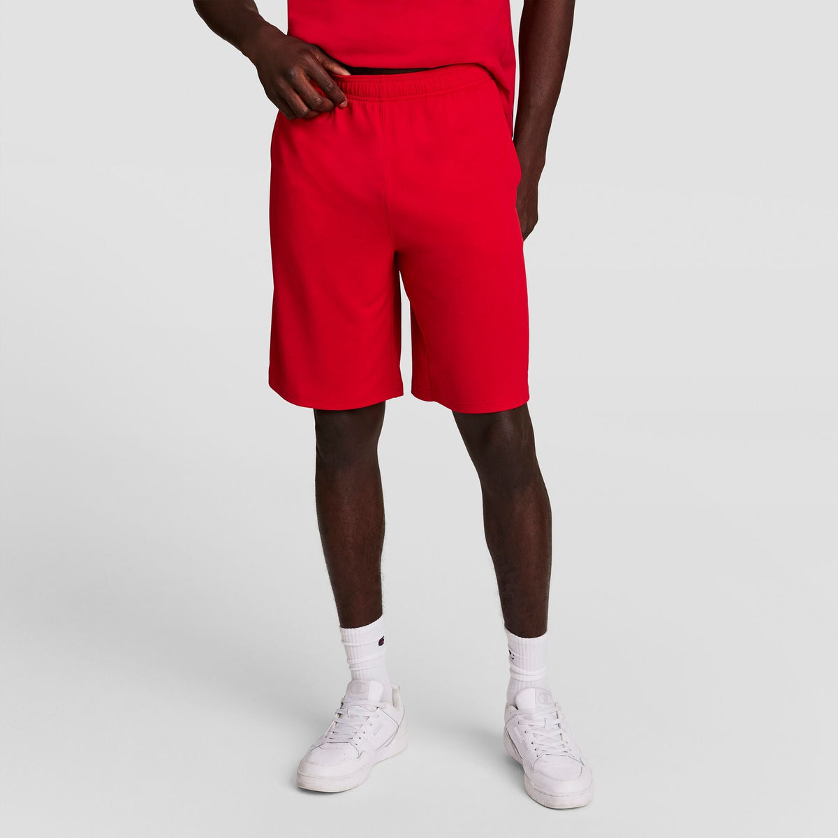 A person poses against a plain background wearing a red T-shirt, Champion 10" Moisture Wicking Sport Shorts, white socks, and white sneakers. The individual’s head is not shown in the image.