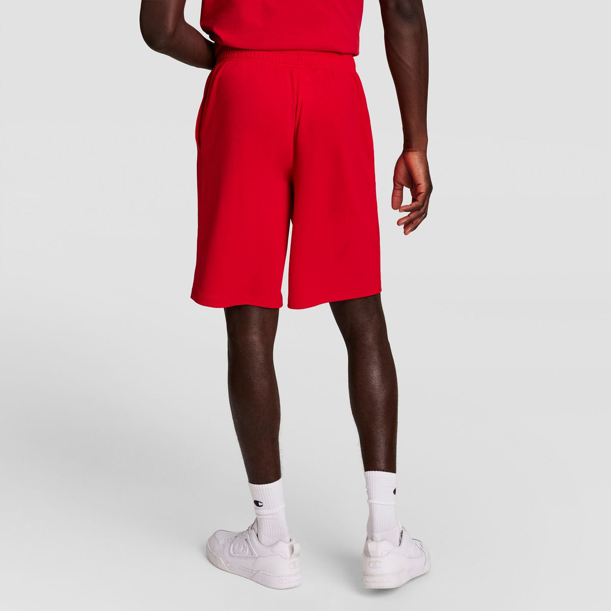 A person stands facing away, wearing Champion 10" Moisture Wicking Sport Shorts in bright red, a matching red shirt, white crew socks, and white sneakers against a plain light gray background.