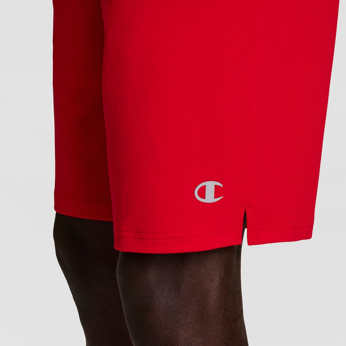 A close-up shows a person wearing Champion 10" Moisture Wicking Sport Shorts in bright red, with the Champion logo near the hem and part of their leg visible against a plain background.