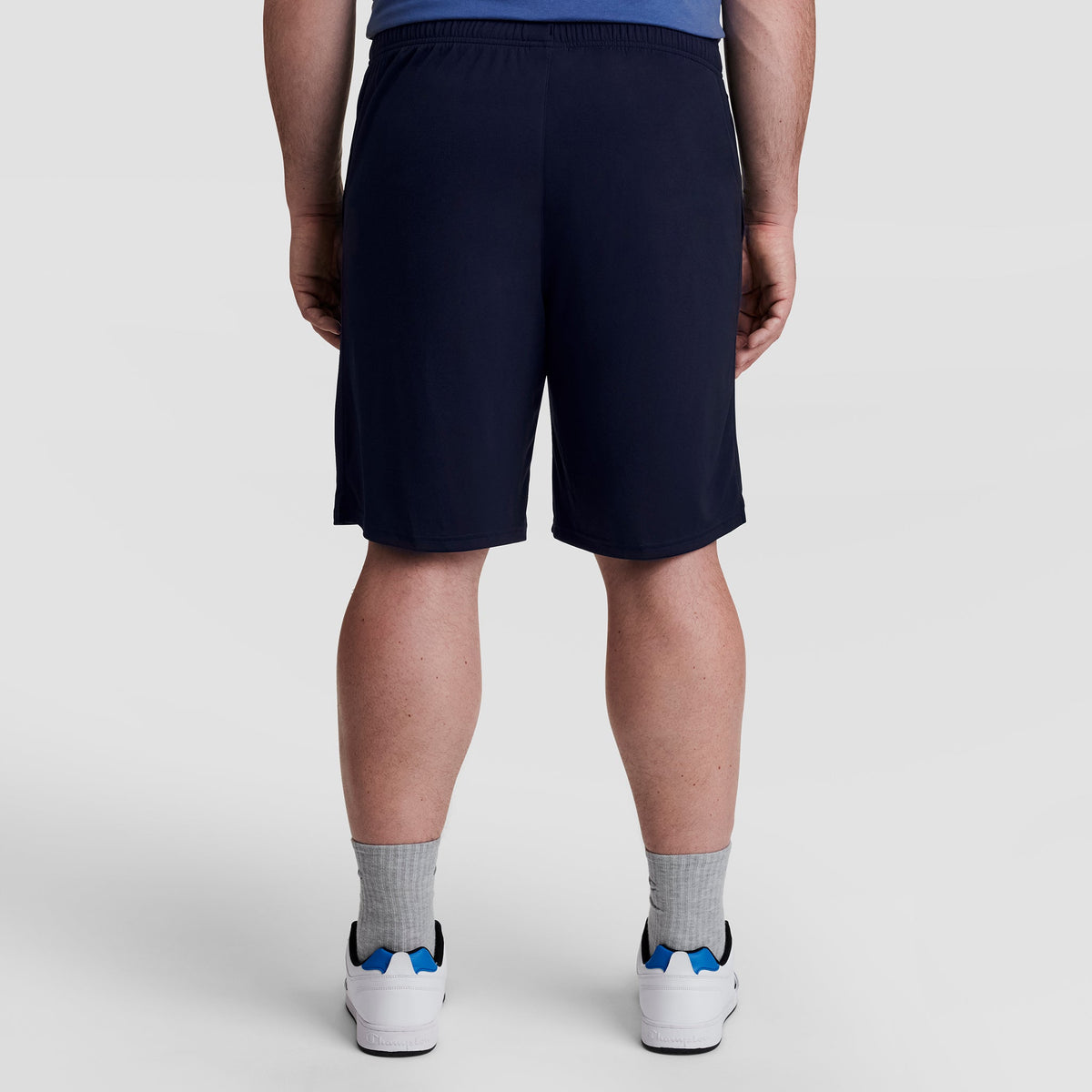 A person stands facing away from the camera, wearing Champion 10" Moisture Wicking Sport Shorts - Big & Tall in navy, gray socks, white sneakers with blue accents, and a blue shirt against a light plain background.