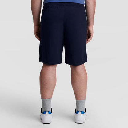 A person stands facing away from the camera, wearing Champion 10" Moisture Wicking Sport Shorts - Big & Tall in navy, gray socks, white sneakers with blue accents, and a blue shirt against a light plain background.