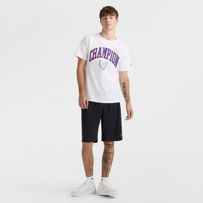 A young man with tattoos stands casually against a white backdrop wearing a white Champion tee, 10" Moisture Wicking Sport Shorts by Champion in black, and white sneakers, with one hand partly in his pocket.