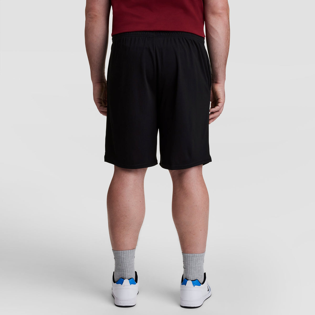 A person stands with their back to the camera, wearing a maroon shirt, gray socks, white sneakers with blue accents, and Champion 10" Moisture Wicking, Sport Shorts - Big & Tall against a plain light background.