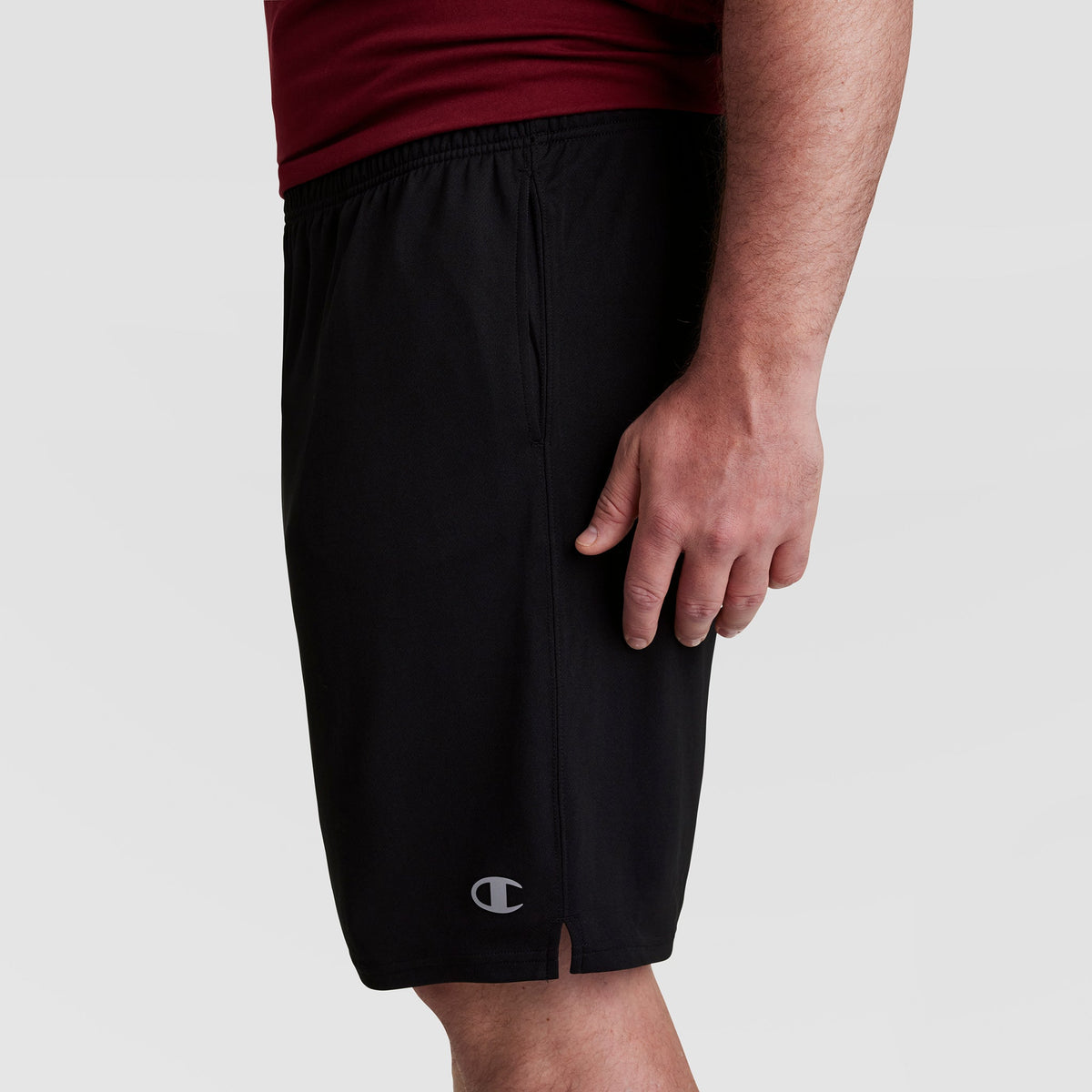 A person wearing Champion 10" Moisture Wicking Sport Shorts - Big & Tall in black and a maroon shirt stands against a plain background, shown from the waist to just above the knee in side profile.