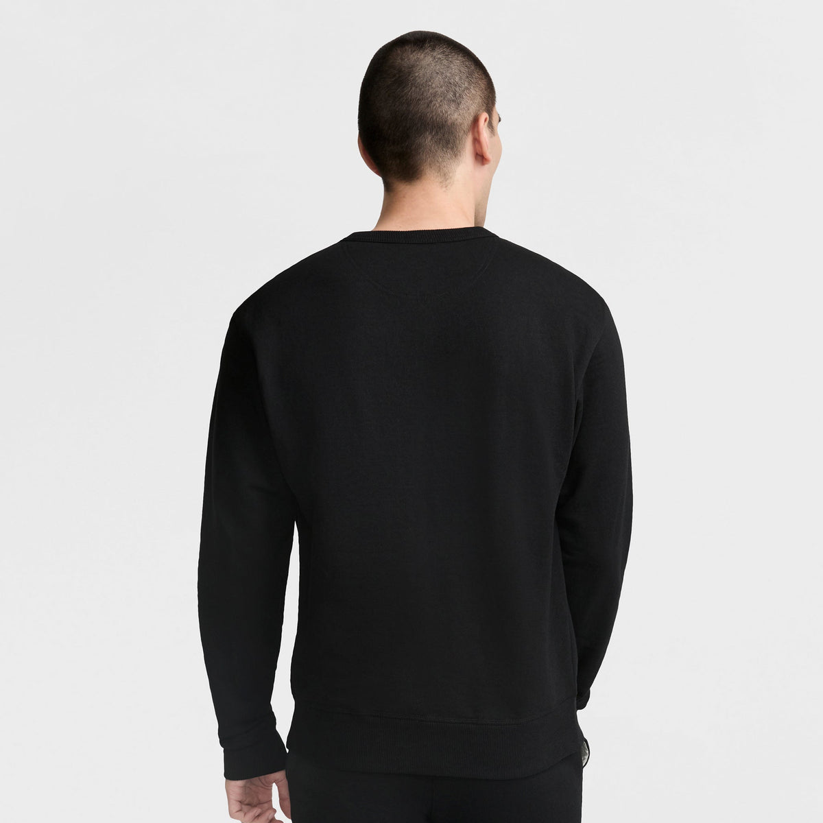 A person with short hair stands facing away from the camera against a plain light background, wearing a black Champion Powerblend Crewneck Sweatshirt with the Classic Script Logo.