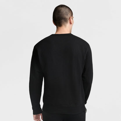 A person with short hair stands facing away from the camera against a plain light background, wearing a black Champion Powerblend Crewneck Sweatshirt with the Classic Script Logo.