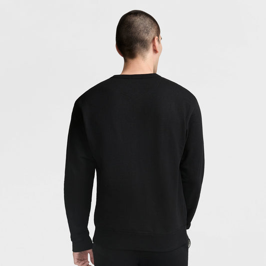 A person with short hair stands facing away from the camera against a plain light background, wearing a black Champion Powerblend Crewneck Sweatshirt with the Classic Script Logo.