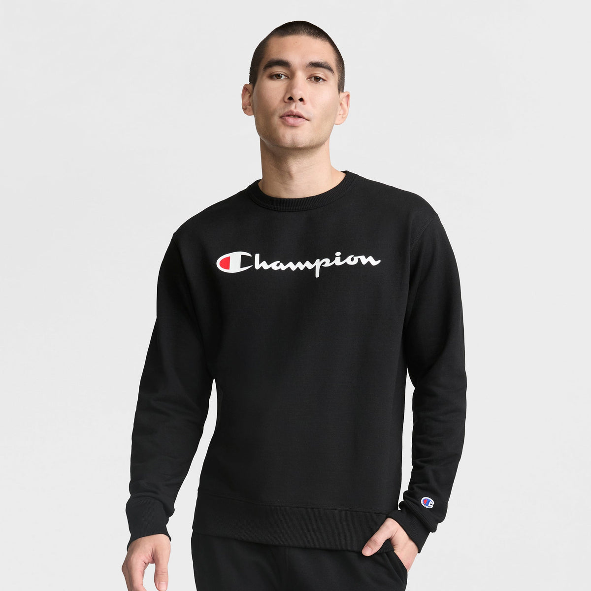 A person stands against a plain background wearing the Champion Powerblend Crewneck Sweatshirt, Classic Script Logo, featuring the Champion logo on chest and sleeve for signature style and comfort.