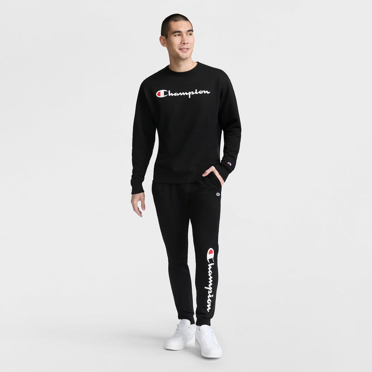 A man stands against a white background wearing a black Champion Powerblend Crewneck Sweatshirt, Classic Script Logo, and matching joggers with white sneakers. The Champion logo is visible on both the sweatshirt and pants.