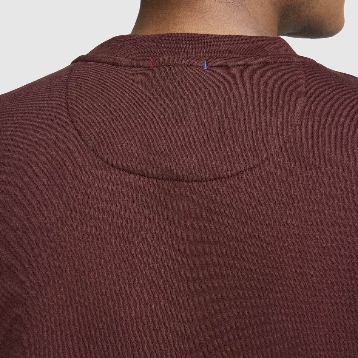 Close-up of the back of a person’s neck and shoulders wearing a maroon Champion Powerblend Crewneck Sweatshirt, Classic Script Logo, featuring visible stitching details and small red and blue threads on the collar.