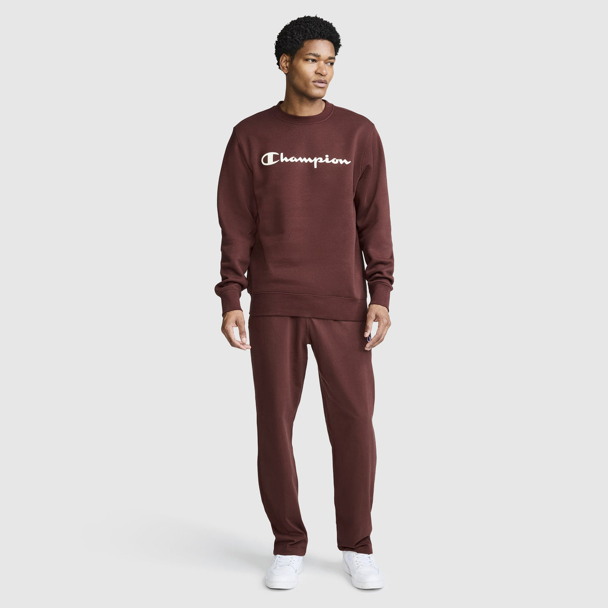A person stands against a plain white background, looking to the side, dressed in a maroon Champion Powerblend Crewneck Sweatshirt with the classic script logo and matching pants. They are also wearing white sneakers, highlighting the comfort and timeless style of this Champion sweatshirt.