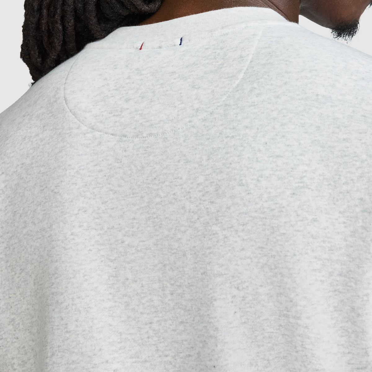 Close-up of the back of a person’s light gray Champion Powerblend Crewneck Sweatshirt, Tonal Script Logo, highlighting the upper back and neck area. A small red, white, and blue stitched Champion detail appears near the neckline. Part of the person’s face and hair is visible.