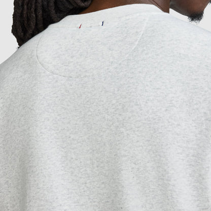 Close-up of the back of a person’s light gray Champion Powerblend Crewneck Sweatshirt, Tonal Script Logo, highlighting the upper back and neck area. A small red, white, and blue stitched Champion detail appears near the neckline. Part of the person’s face and hair is visible.