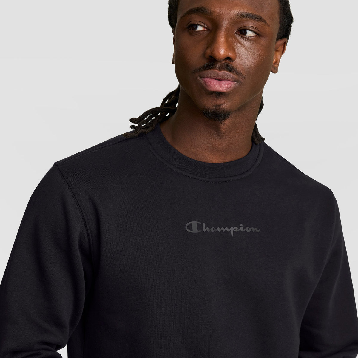 A man with long braids models the Champion Powerblend Crewneck Sweatshirt with a tonal script logo in black, gazing slightly to the side against a plain light gray background.