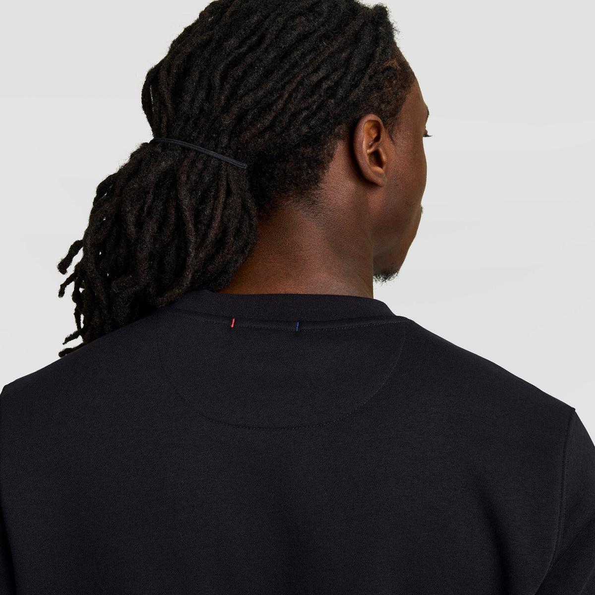 A person with long, dark dreadlocks tied back is seen from behind wearing a black Champion Powerblend Crewneck Sweatshirt, Tonal Script Logo, against a plain light background.