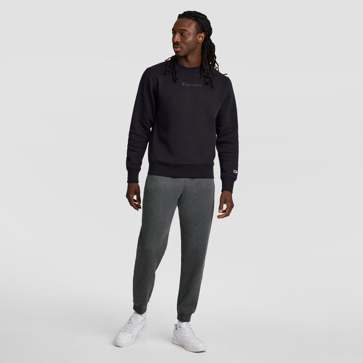 A person stands against a light background wearing the Champion Powerblend Crewneck Sweatshirt with Tonal Script Logo in black, paired with gray sweatpants and white sneakers, looking slightly to the side with one hand down.