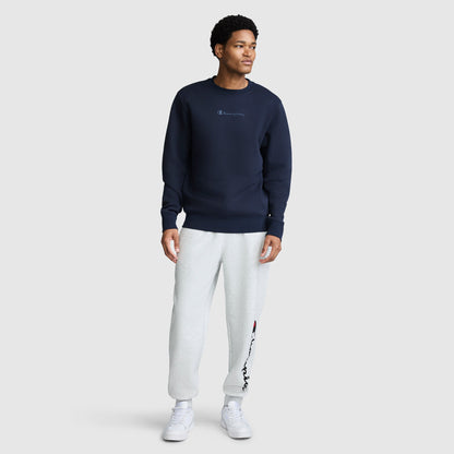A man stands against a plain light gray background wearing a Champion Powerblend Crewneck Sweatshirt with a tonal script logo in navy blue, paired with light gray sweatpants featuring a logo on the left leg, and white sneakers.