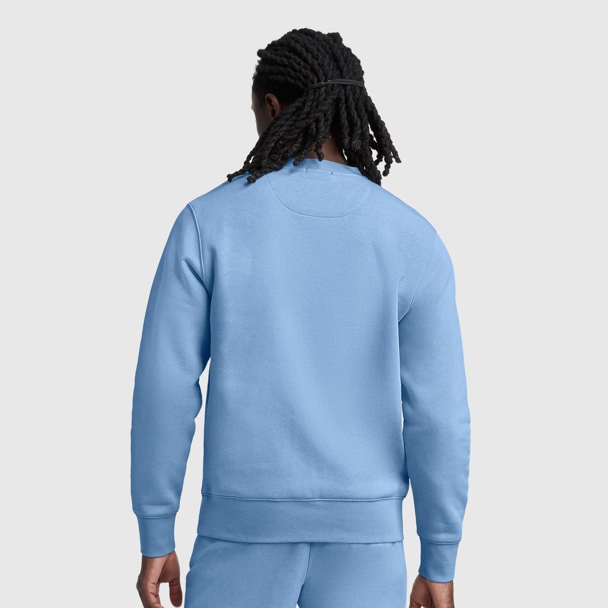 A person with long, dark, braided hair is shown from the back, wearing a light blue Champion Powerblend Crewneck Sweatshirt with a tonal script logo and matching sweatpants against a plain, light gray background.