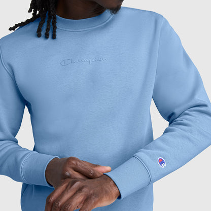 A person wearing a light blue Champion Powerblend Crewneck Sweatshirt, Tonal Script Logo, with subtle embroidered branding on the chest and sleeve, adjusts their sleeve while facing slightly away from the camera.