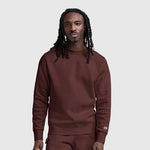 A man with long dreadlocks wears a maroon Champion Powerblend Crewneck Sweatshirt with a tonal script logo and matching pants, standing against a plain light gray background.
