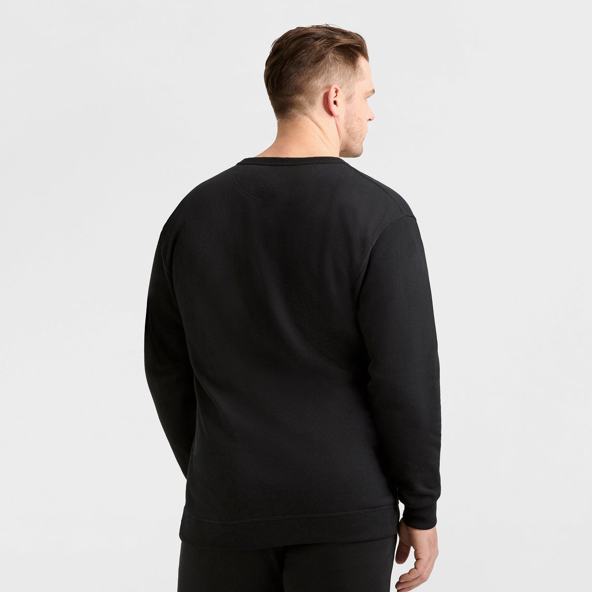 A man with short hair, facing away from the camera, is wearing a plain black Champion Powerblend Crewneck Sweatshirt, C Logo - Big & Tall, and black pants against a light gray background.
