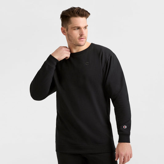 A man models the Champion Powerblend Crewneck Sweatshirt, C Logo – Big & Tall, in plain black with small logos on the sleeve and chest, standing against a white background and looking to his left with one hand near his neck.