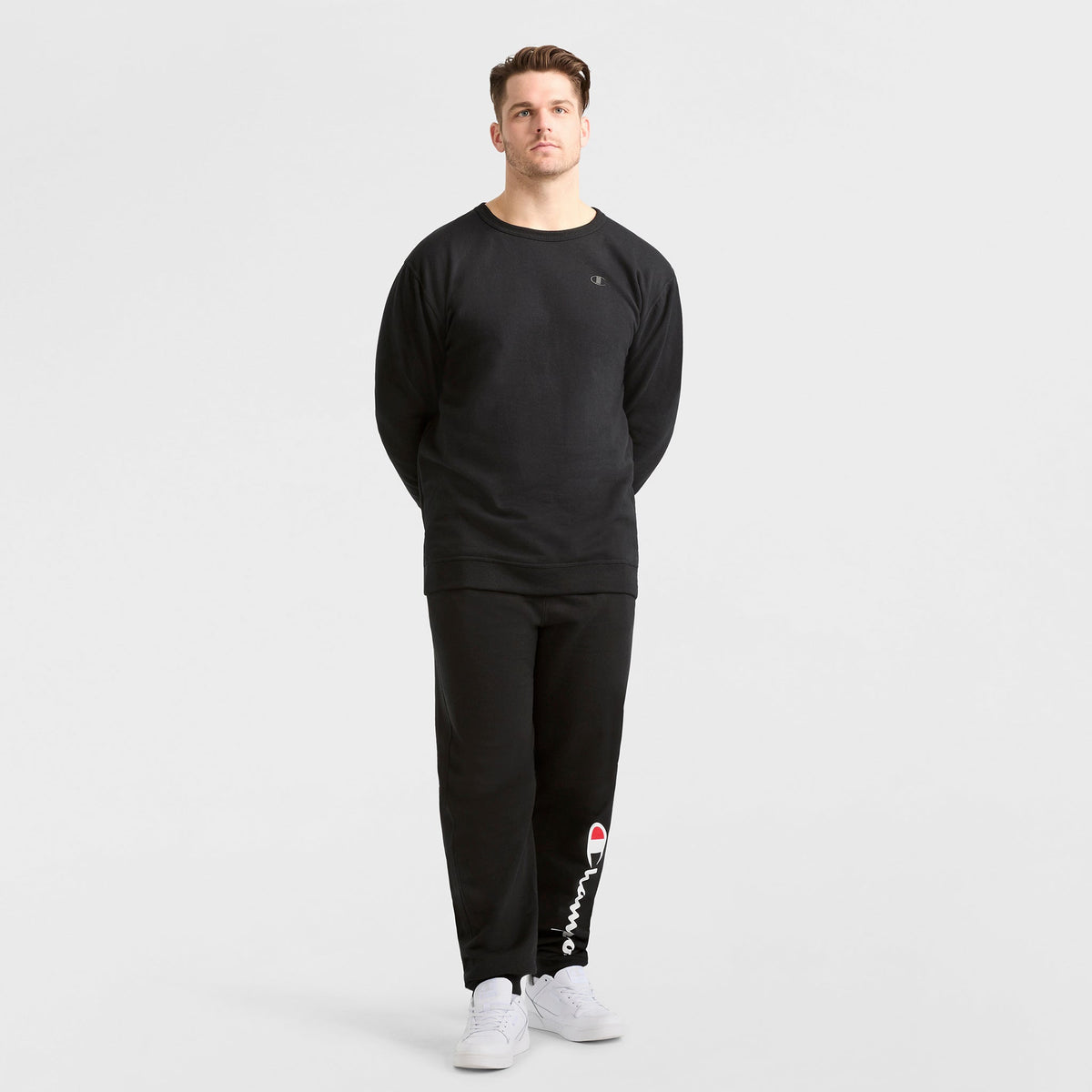 A man stands against a plain white background wearing a Champion Powerblend Crewneck Sweatshirt, C Logo - Big & Tall, and matching Champion sweatpants with white sneakers. His hands are behind his back.