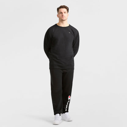 A man stands against a plain white background wearing a Champion Powerblend Crewneck Sweatshirt, C Logo - Big & Tall, and matching Champion sweatpants with white sneakers. His hands are behind his back.