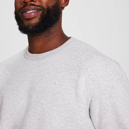 A bearded man smiles subtly, wearing a light grey Champion Powerblend Crewneck Sweatshirt, C Logo - Big & Tall. Only his lower face and upper torso are shown against a plain white background.
