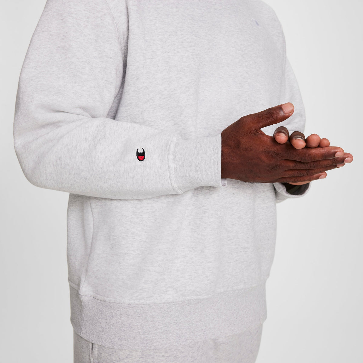 A person wears a light gray Champion Powerblend Crewneck Sweatshirt, C Logo - Big & Tall, with matching pants. A small embroidered red and white cartoon face decorates the sleeve near the wrist.