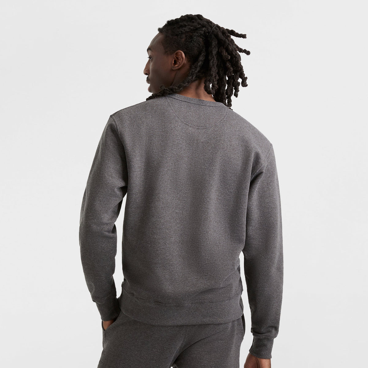 Seen from behind, a person with long, twisted hair wears a dark gray Champion Powerblend Crewneck Sweatshirt (C Logo) and matching pants against a plain white background.
