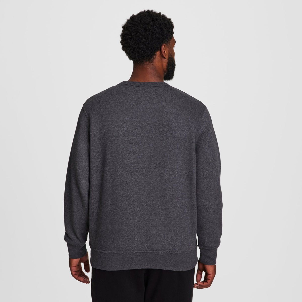 A man with short curly hair faces away from the camera, wearing a dark gray Champion Powerblend Crewneck Sweatshirt, C Logo - Big & Tall, and black pants against a plain white background.