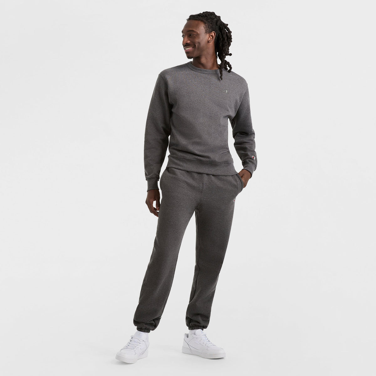 Wearing a matching dark gray Champion Powerblend Crewneck Sweatshirt with the C Logo and sweatpants, paired with white sneakers, a person stands against a plain white background, looking to the side and smiling.