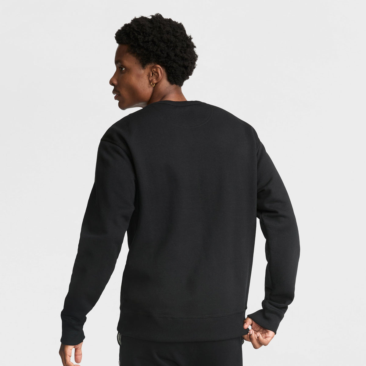 A person with short, curly hair is seen from behind in a plain black Champion Powerblend Crewneck Sweatshirt, C Logo, and black pants, standing against a white background.