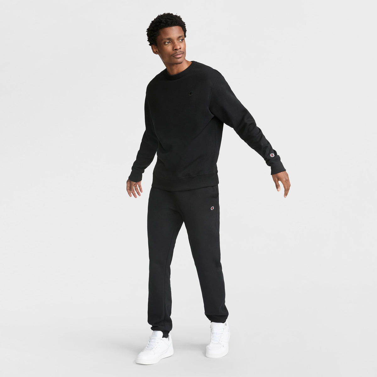 A man stands against a plain white background wearing a Champion Powerblend Crewneck Sweatshirt with C Logo, matching black sweatpants, and white sneakers. He faces slightly to the side with relaxed arms and a neutral expression.