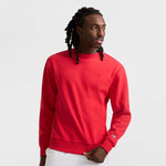 A man with long braided hair is wearing a bright red Champion Powerblend Crewneck Sweatshirt with the C Logo and white pants, standing against a plain white background while looking to his left.