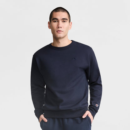 A man wears a Champion Powerblend Crewneck Sweatshirt, C Logo, paired with navy blue pants, standing with hands in his pockets against a plain white background.